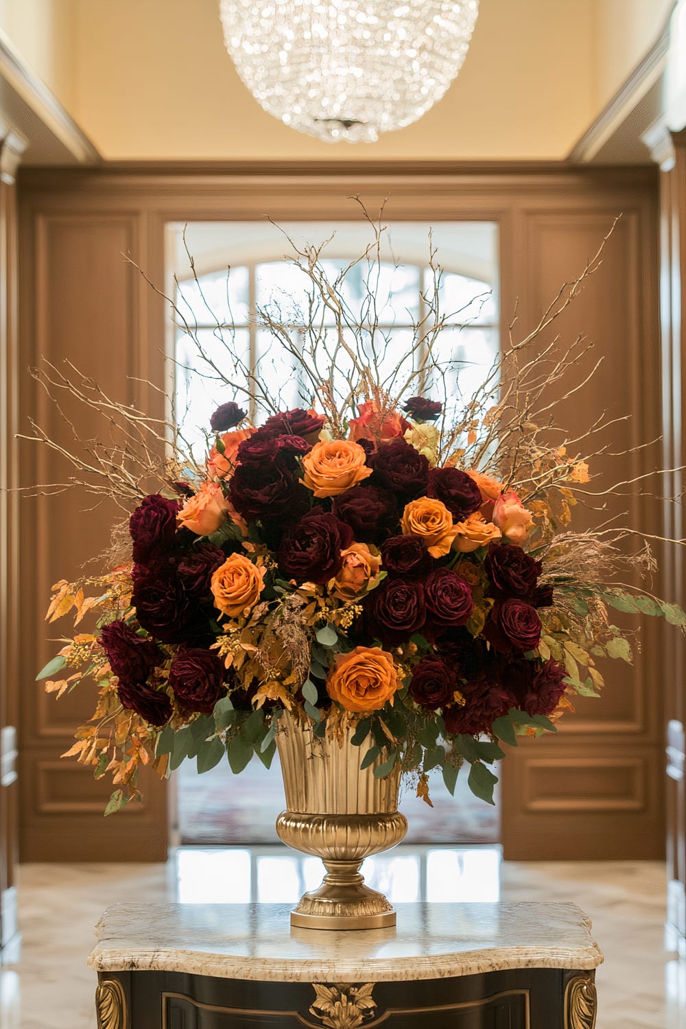 An elegant floral arrangement featuring deep burgundy and vibrant orange roses is displayed in a gold vase on a decorative marble-topped table. The arrangement includes dried branches and additional greenery. It is set against a luxurious interior backdrop, featuring wooden paneling and a grand chandelier hanging from the ceiling.