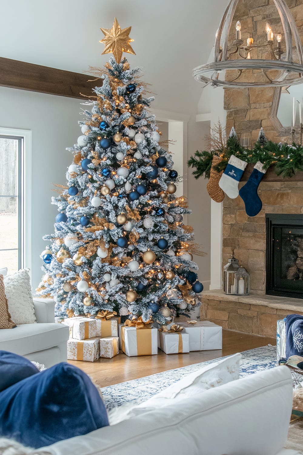 A well-decorated living room featuring a Christmas tree adorned with blue, gold, and white ornaments, and golden leaves. At its top is a golden star tree topper. Wrapped gifts with gold ribbons sit beneath the tree. Adjacent to the tree, a fireplace surrounded by a rustic stone mantle is decorated with garland and stocking hangers. A large, elegant chandelier with exposed bulbs hangs above the scene. The room includes a white sofa with blue and white pillows, and a window through which the outdoor scenery is visible.