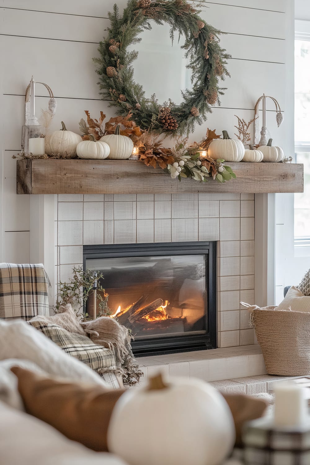 An elegantly decorated living room features a white-glazed fireplace with a rustic wooden mantel. Atop the mantel are white decorative pumpkins, fall foliage, and lanterns, adding a touch of autumn charm. Above the mantel hangs a large wreath made of evergreen sprigs and pine cones. A plaid throw blanket and pillows with soft, earthy tones can be seen in the foreground, creating a warm and inviting atmosphere.