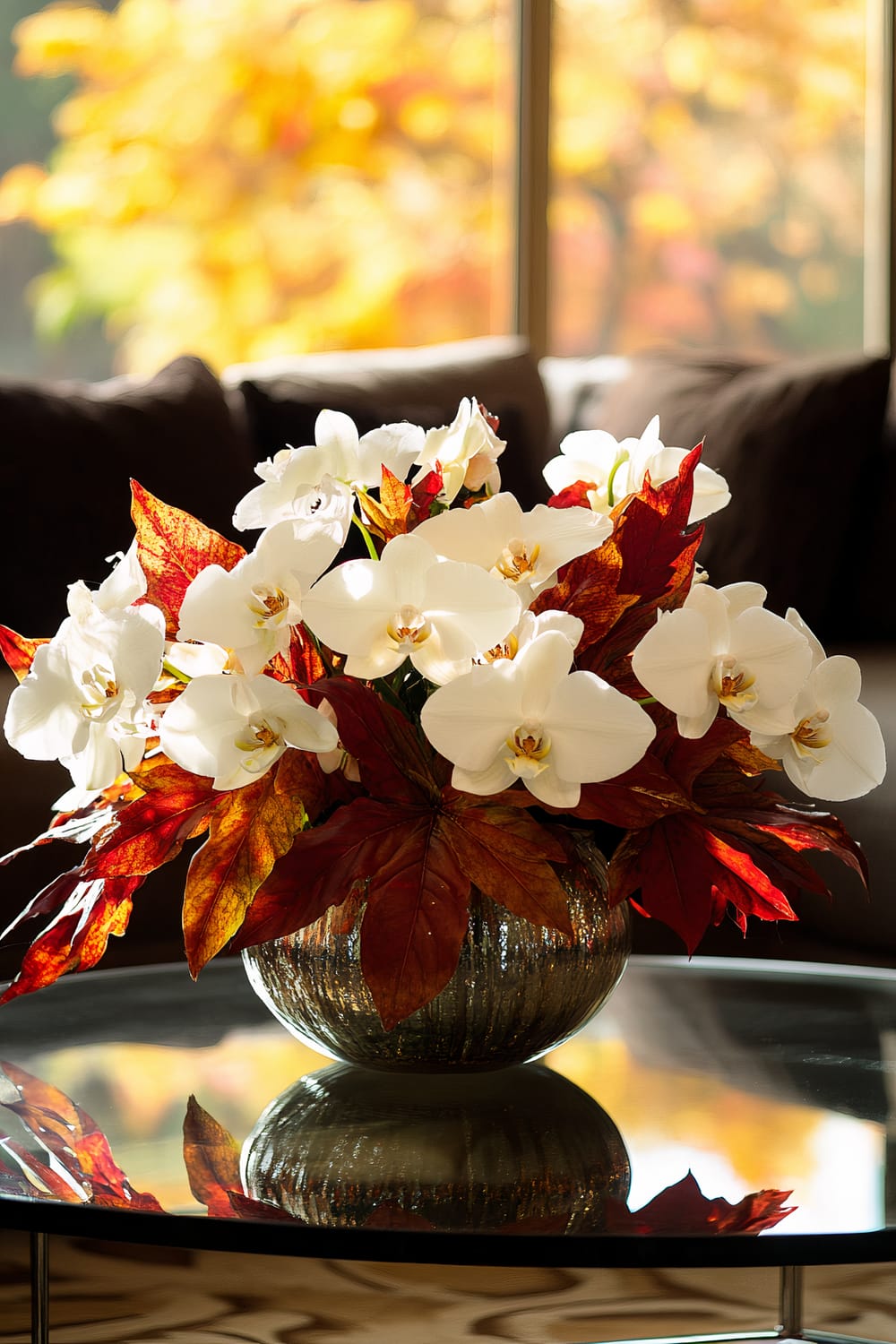 A glass coffee table is adorned with a bold arrangement of white orchids set among red and orange leaves. The white flowers stand out prominently against the fall-colored foliage, and the arrangement is contained in a reflective, textured glass bowl. In the background, there is a blurred autumnal scene with warm hues of yellow and orange.