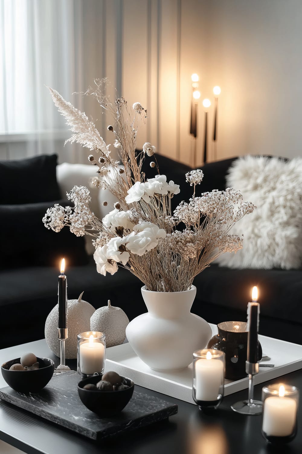 An elegant monochrome Thanksgiving coffee table setup featuring a sleek black and white color scheme. The table is adorned with a white ceramic vase holding white and black flowers, flanked by black candle holders with burning candles. Surrounding the table are gray and white cushions on a modern black sofa. The setting has soft ambient lighting, creating a sophisticated and uncluttered look.