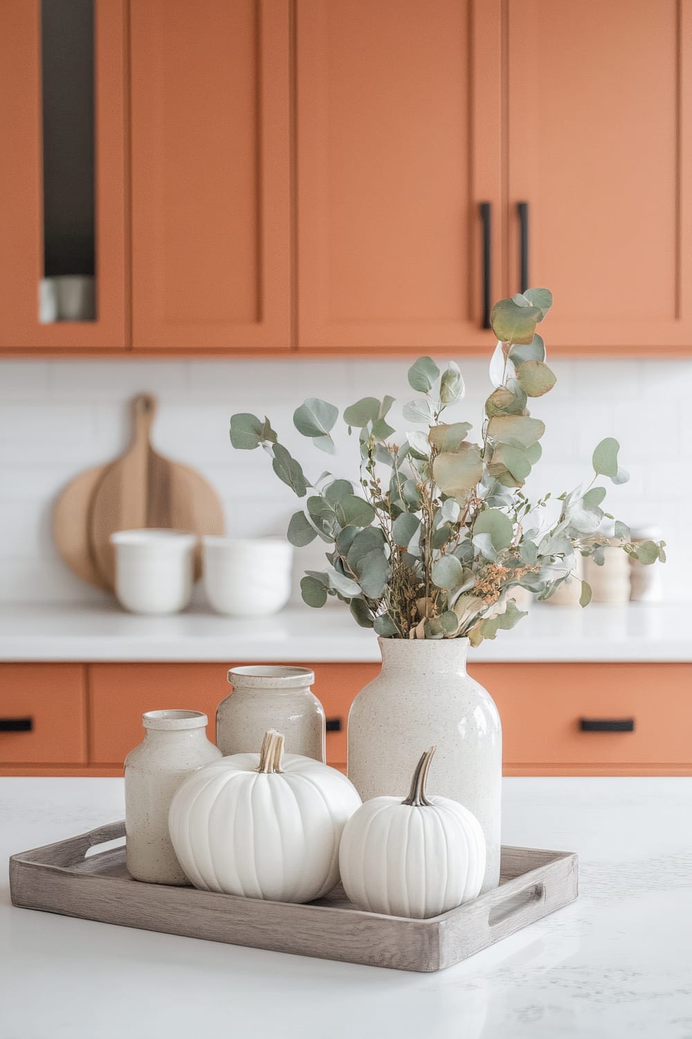 A well-designed kitchen counter featuring a rustic arrangement consisting of eucalyptus leaves in a ceramic vase, surrounded by smaller vases and decorative white pumpkins, all placed on a wooden tray. The background showcases orange cabinets with black handles, a white tiled backsplash, and additional kitchen items, including wooden cutting boards and white ceramic containers.