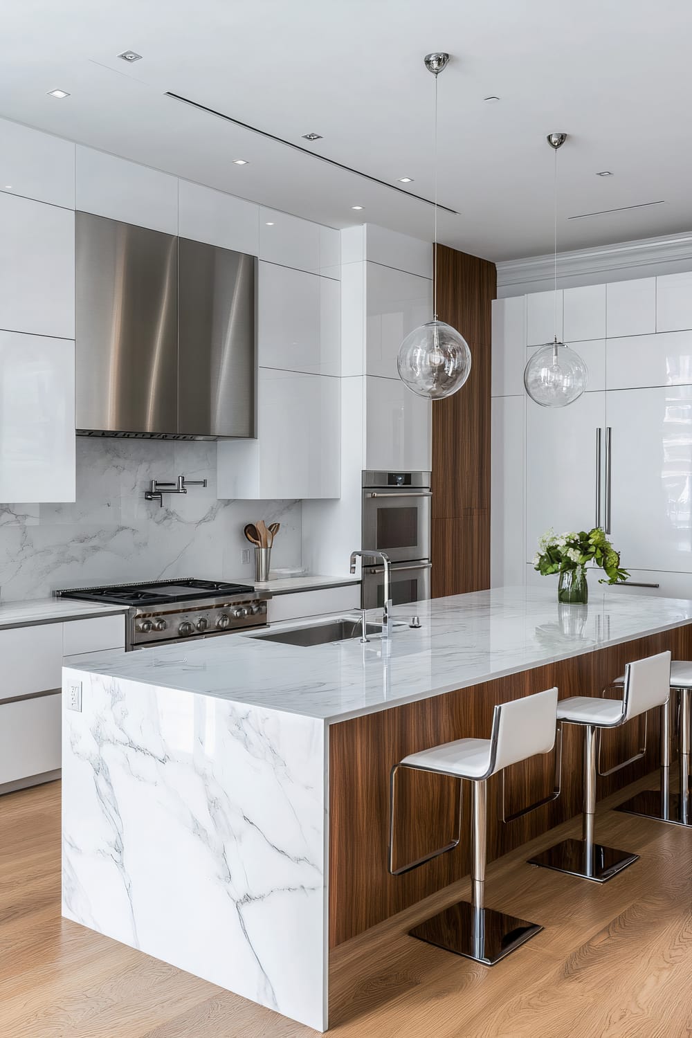 A modern kitchen featuring a large island with a white marble countertop and wood paneling, accompanied by three white bar stools. The island includes a built-in sink and a green plant in a glass vase. The kitchen backdrop showcases high-gloss white cabinetry, a stainless steel stove, and a double oven, with a marble backsplash. Two spherical glass pendant lights hang above the island, and the kitchen floor is covered with light brown wood.