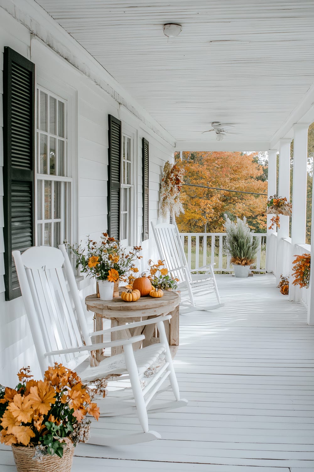 A white wooden porch adorned with fall-themed decorations. Two white rocking chairs are positioned around a small wooden table which holds yellow flowers and mini pumpkins. The porch railings have similar fall decorations. Greenery and fall foliage can be seen in the background.