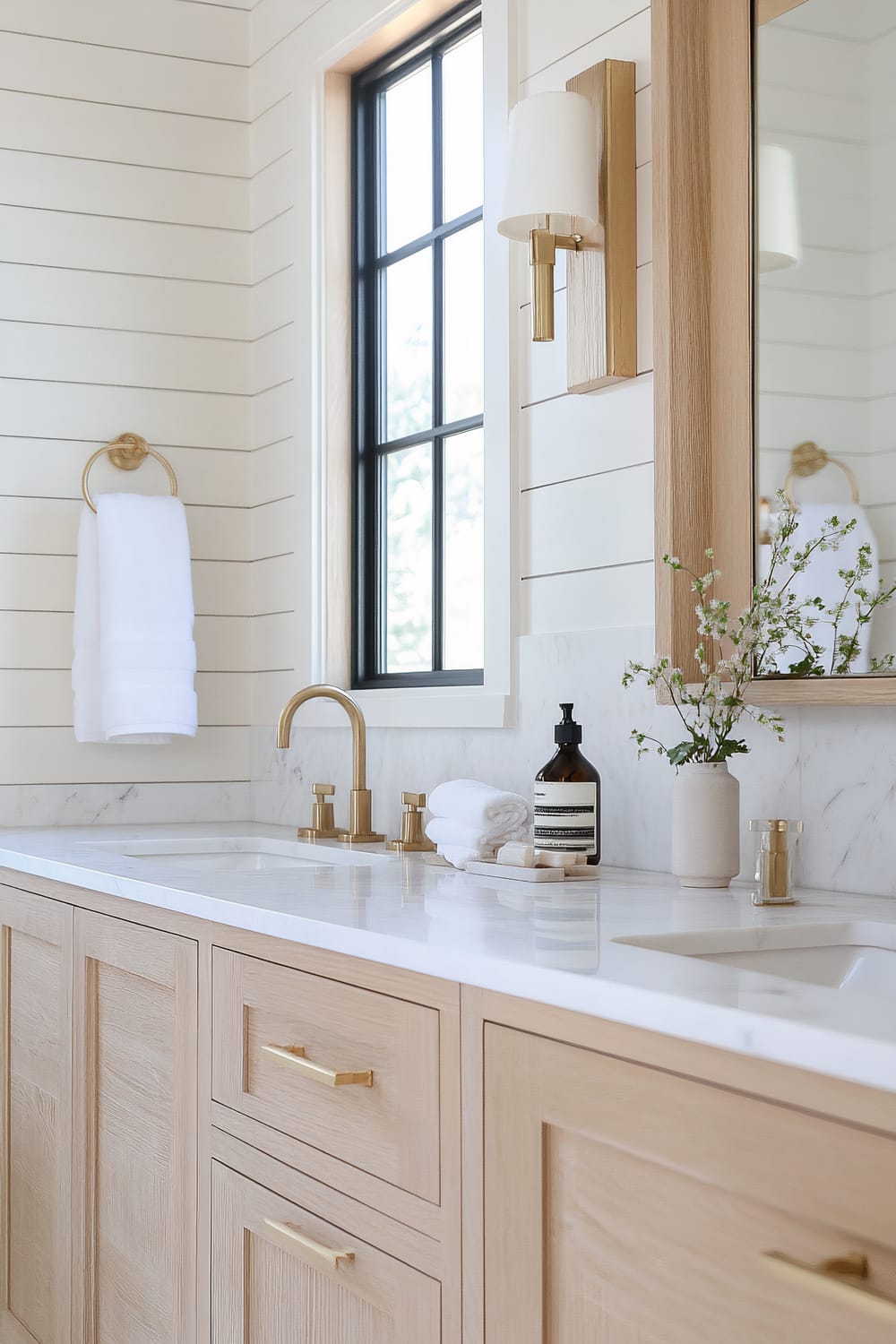 This image shows a modern bathroom featuring a double-sink vanity. The cabinets are made of light wood with minimalist gold handles. The countertop is white marble with subtle gray veining. Mounted above each sink are gold faucet sets. A large rectangular mirror framed in light wood hangs above the sinks. To the right of the mirror, a modern light fixture with a white lampshade and gold base is mounted on the wall. On the countertop, there are neatly folded white towels, a soap dispenser, and a small vase holding delicate green and white flowers. A large window framed in black metal trim provides natural light and view of greenery outside. A white towel hangs on a gold towel ring to the left of the sink.