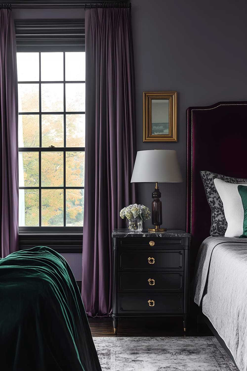 A stylish bedroom featuring a large window with royal purple satin drapes, an oxblood red vintage wood headboard, a gray duvet with emerald green accents, a matte black marble nightstand with a minimalist lamp, and a gold leafed brass mirror hanging above the bed reflecting the room’s décor.