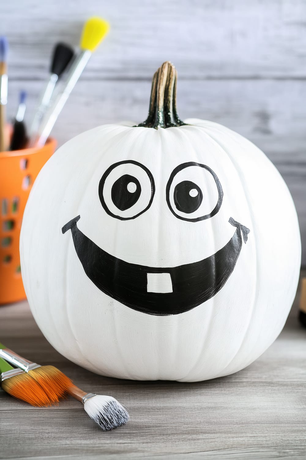 A white pumpkin with a large painted smiling face, featuring two large, circular eyes and a big black smile with one tooth. The pumpkin's surface is painted entirely white, while the facial features are painted black. In the background, there are several paintbrushes of various sizes in an orange container, and more paintbrushes are laid on the wooden surface near the pumpkin.