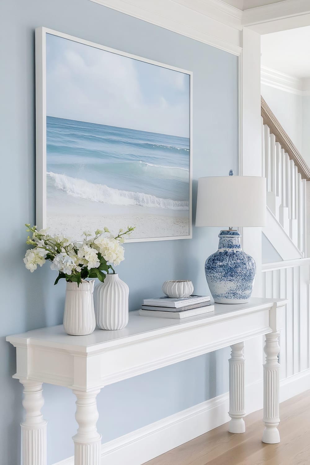 A tastefully decorated interior space features a white console table against a light blue wall. Above the table hangs a large, serene seascape painting that showcases calm blue waters and gently breaking waves, framed in white. The table is topped with two white ceramic vases filled with white flowers, a stack of books adorned with small decorative items, and a prominent blue and white ceramic lamp with a white lampshade. The background reveals a staircase with white spindles and a wood banister.