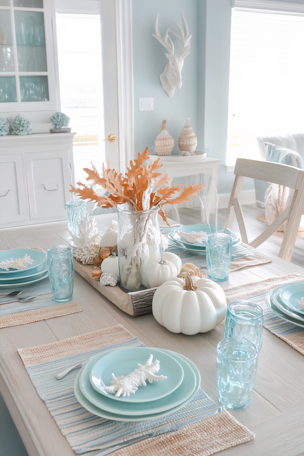 A serene dining area with a coastal theme is depicted. The dining table is adorned with a centerpiece featuring white pumpkins, orange leaves, and white corals in a glass vase, all arranged on a rustic wooden tray. The table setting includes light blue plates, clear blue glasses, and woven placemats in muted tones of beige and blue. Behind the table, the room features a white sideboard with glass-fronted cabinets displaying a collection of blue glassware, topped with nautical decor items. The walls are painted in a soft seafoam green, and a white stag head wall decor adds a whimsical touch. A glimpse of a cozy sitting area with light-colored furniture can be seen in the background.