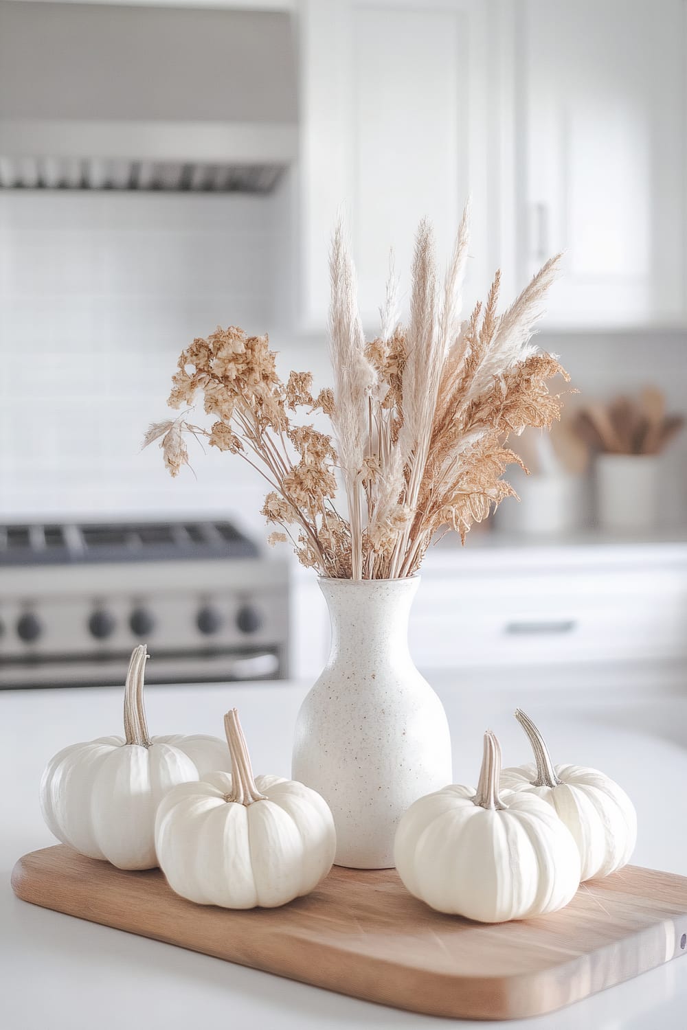 An elegantly minimalist kitchen scene featuring a white ceramic vase with dried floral arrangements placed on a light wooden board. Surrounding the vase are four small white pumpkins. The background shows a sleek, stainless steel stovetop and white cabinets, emphasizing a clean, modern aesthetic.