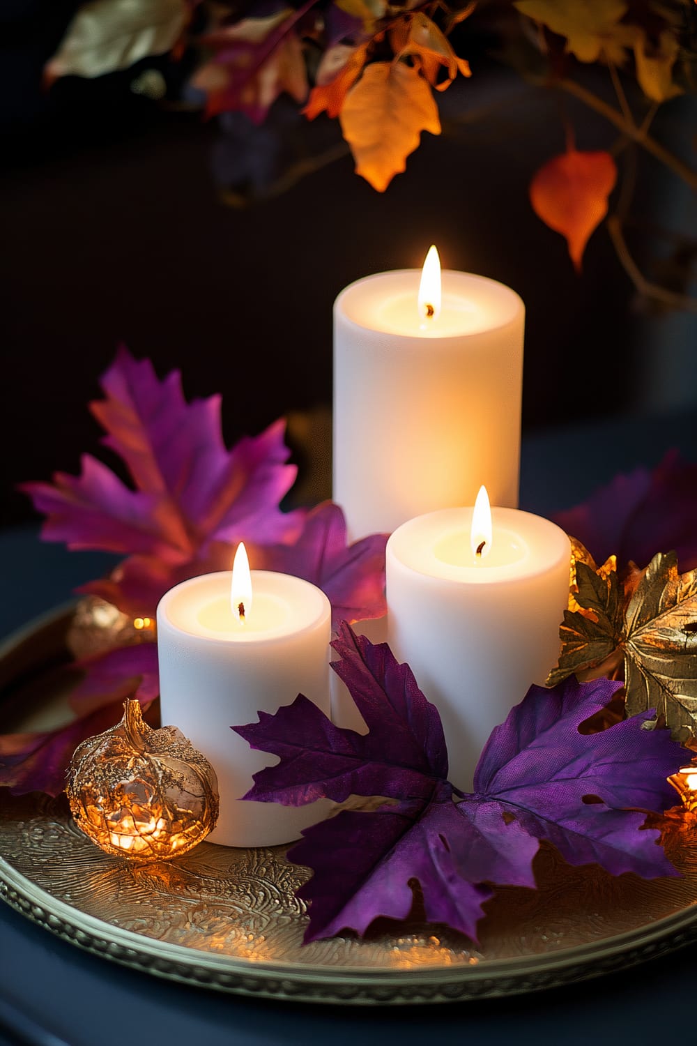 Three tall white candles adorned with vibrant purple and gold artificial fall leaves, placed on an ornate gold tray. One small decorative item resembling a pumpkin is also part of the arrangement, illuminated by the gentle glow of the candle flames under ambient lighting.