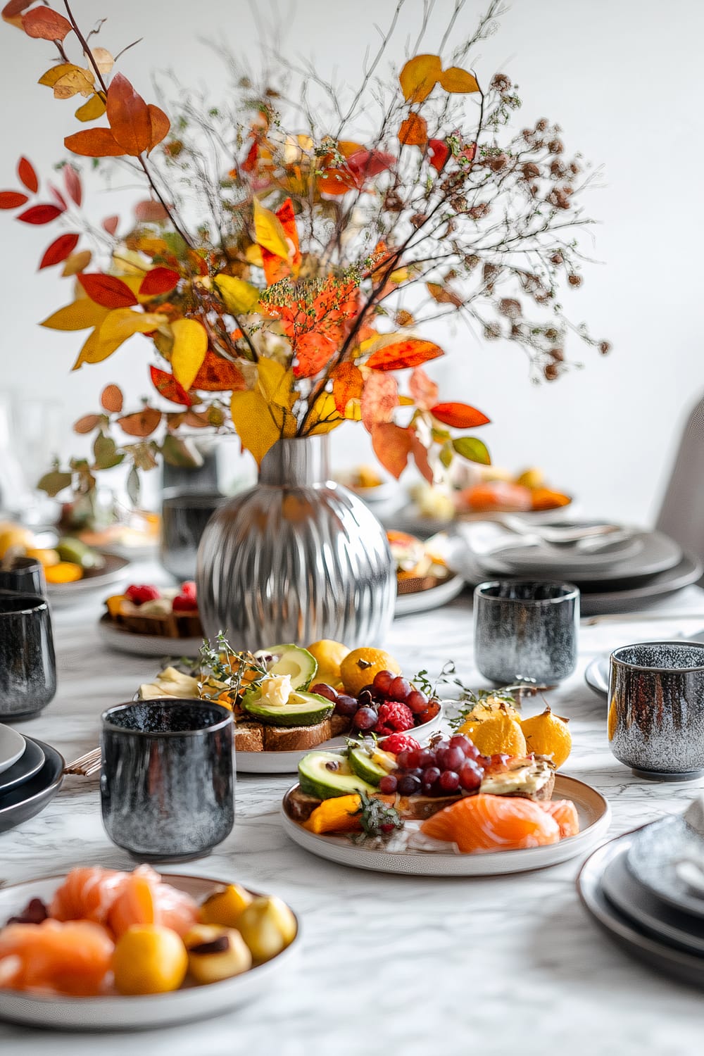 A minimalist Friendsgiving brunch table set on a white marble surface with earth-toned ceramic plates and cobalt blue glass mugs. The plates contain avocado toast, smoked salmon, and fresh fruit salad. The centerpiece is a geometric metallic vase in deep yellow with red autumn leaves and small branches. Another silver metallic vase filled with fresh greenery sits beside the centerpiece against a white backdrop with subtle autumn foliage.