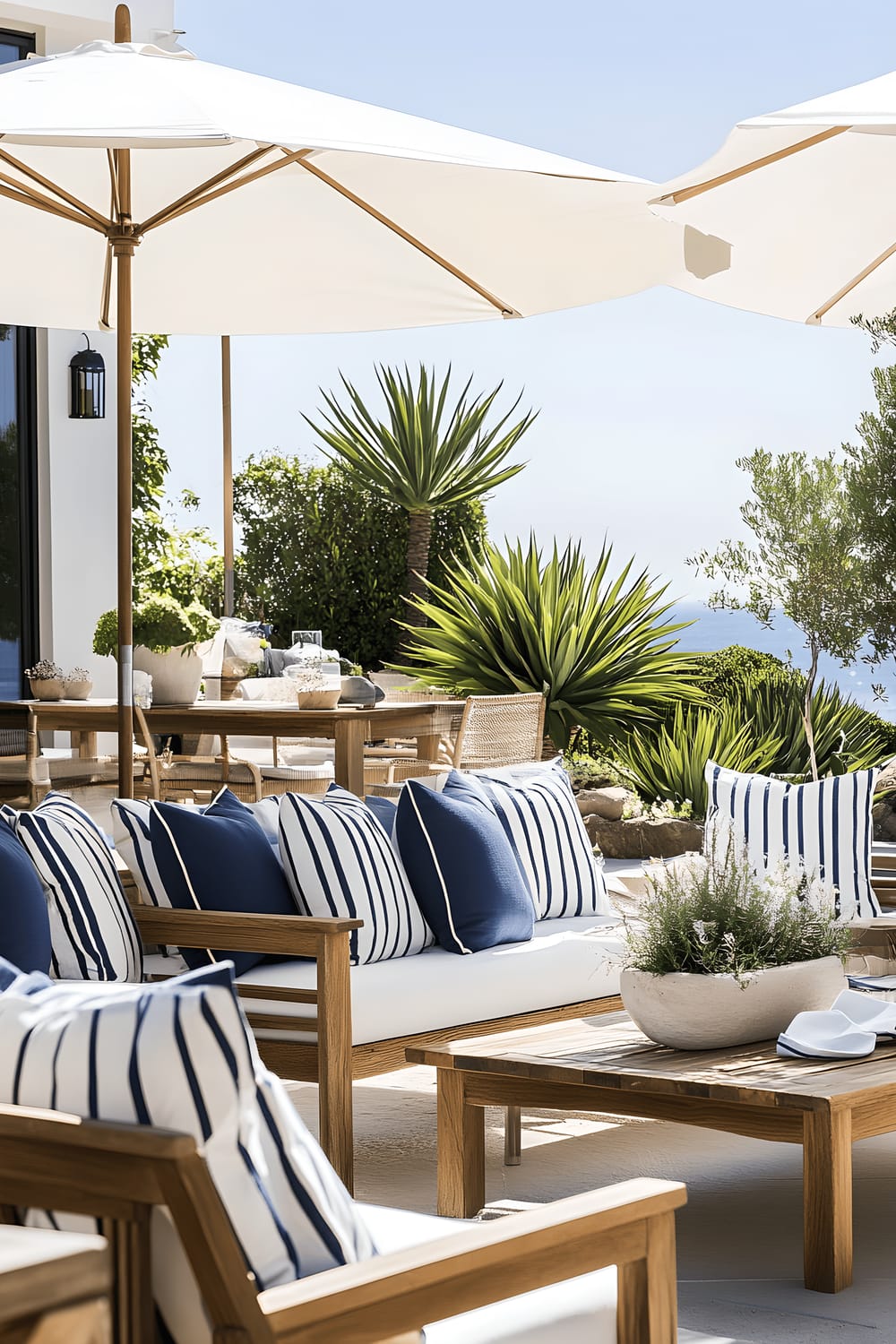 A sunny, relaxed patio featuring a weathered wooden table and chairs set with navy and white striped cushions. The patio is decorated with potted eucalyptus plants and native Australian wildflowers, including kangaroo paw. Large outdoor umbrellas cast shade over the furniture, enhancing the breezy, coastal ambiance.