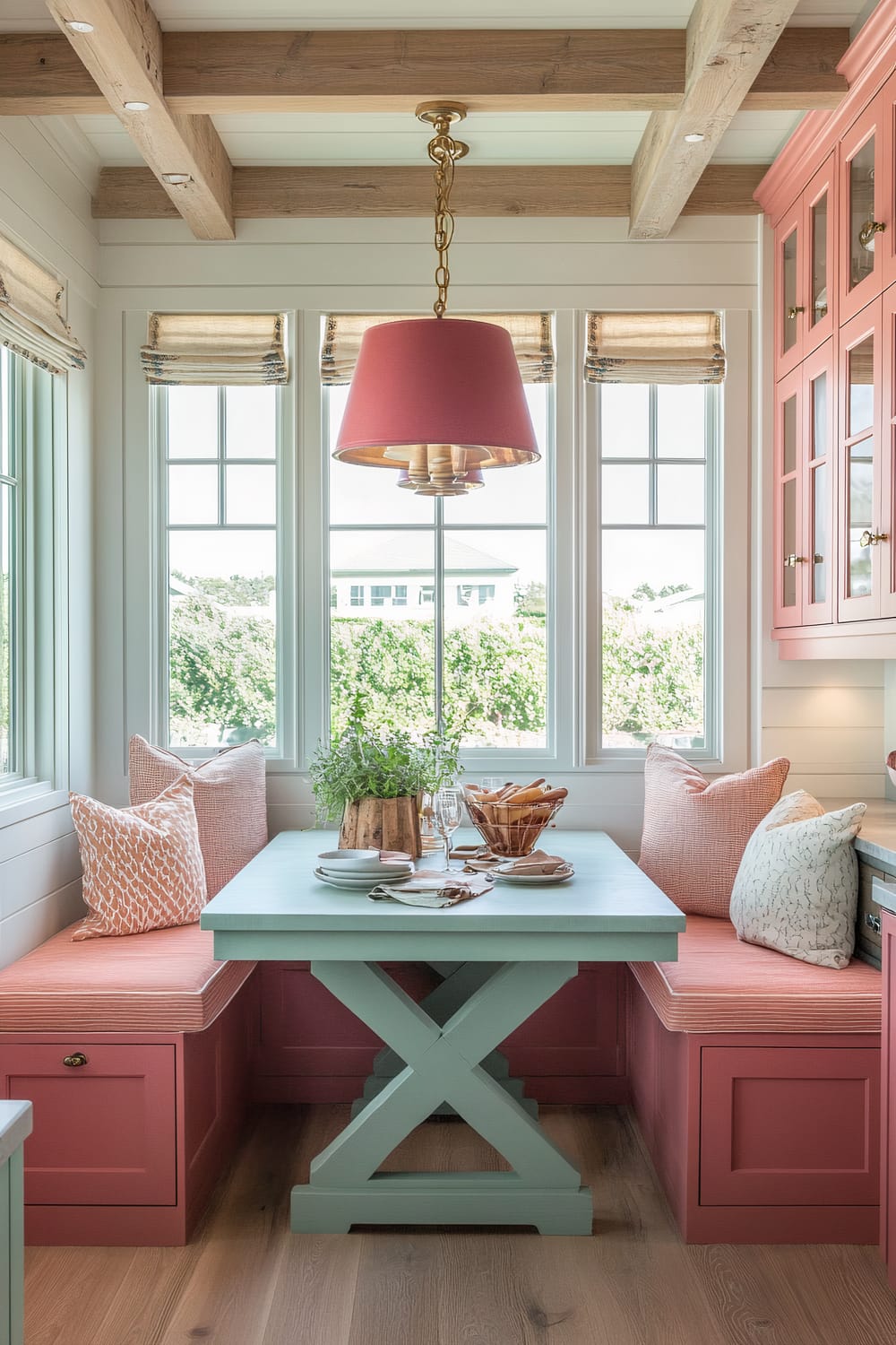 A kitchen nook with a pastel color scheme. The built-in seating features pink cushions and pillows with various patterns. A light blue table with an X-base stands in the center. Above the table hangs a chandelier with a pink lampshade. Large windows provide a view of greenery outside.