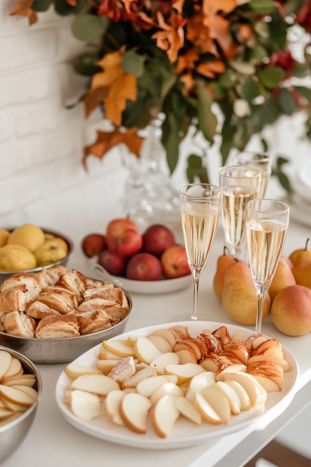 A minimalist Friendsgiving brunch setup featuring a white ceramic platter with assorted sliced pears and apples, surrounded by four metallic bowls of pastries, pears, and apples. Five elegant vintage glasses of mimosa are arranged on a sleek white table, with subtle autumn foliage in a vase in the background.