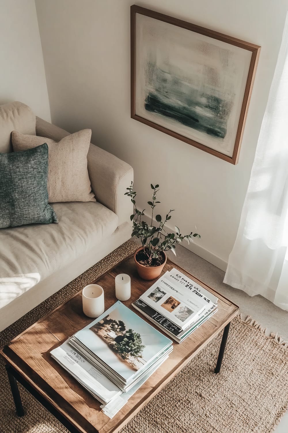 Top-down view of a small living room with a slim wooden coffee table. The table features a stack of magazines, a decorative candle, and a small potted plant. A single piece of abstract art hangs above the table, and natural light from a window illuminates the clean and organized decor. A beige sofa with two pillows is partially visible on the left side of the image.