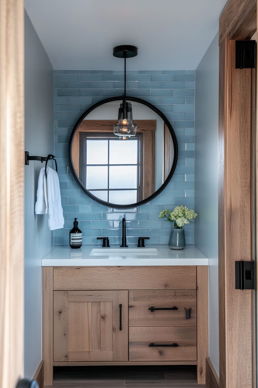 An elegant bathroom features a combination of modern and rustic design elements. A wooden vanity with black handles is topped with a white countertop and black sink fixtures. Above the vanity, a large round mirror with a black frame reflects light, and a single pendant light hangs from the ceiling. The wall behind the vanity is lined with pale blue subway tiles. To the right of the sink, a vase with white flowers adds a touch of nature. A window with wooden trim and frosted glass is visible through the mirror.