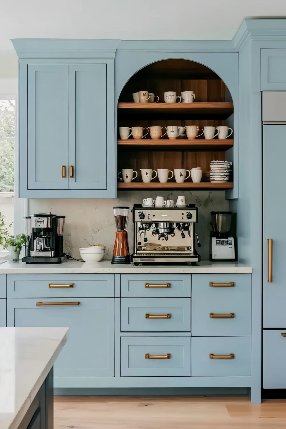 A well-organized kitchen coffee station features light blue cabinetry with brass handles. The upper cabinets have an open arched shelf displaying an assortment of mugs, some of which are plain and others with patterns. Below the shelf is a countertop equipped with a shiny espresso machine, a coffee grinder with an orange base, and a drip coffee maker. The backdrop is a white marble-like backsplash, complementing the overall design.