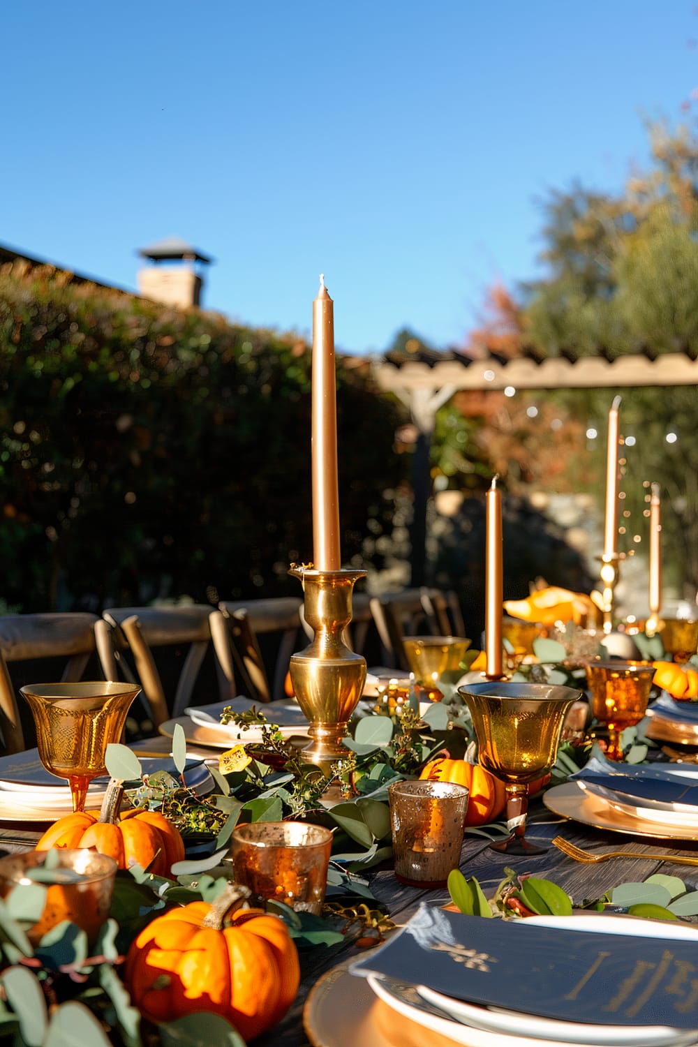 An elegantly decorated outdoor dining table is set under a clear blue sky. The table features brass candle holders with long, thin candles, amber glasses, small pumpkins, and green foliage. Plates and menus are neatly arranged in a sophisticated fashion, reflecting a festive and refined setting.