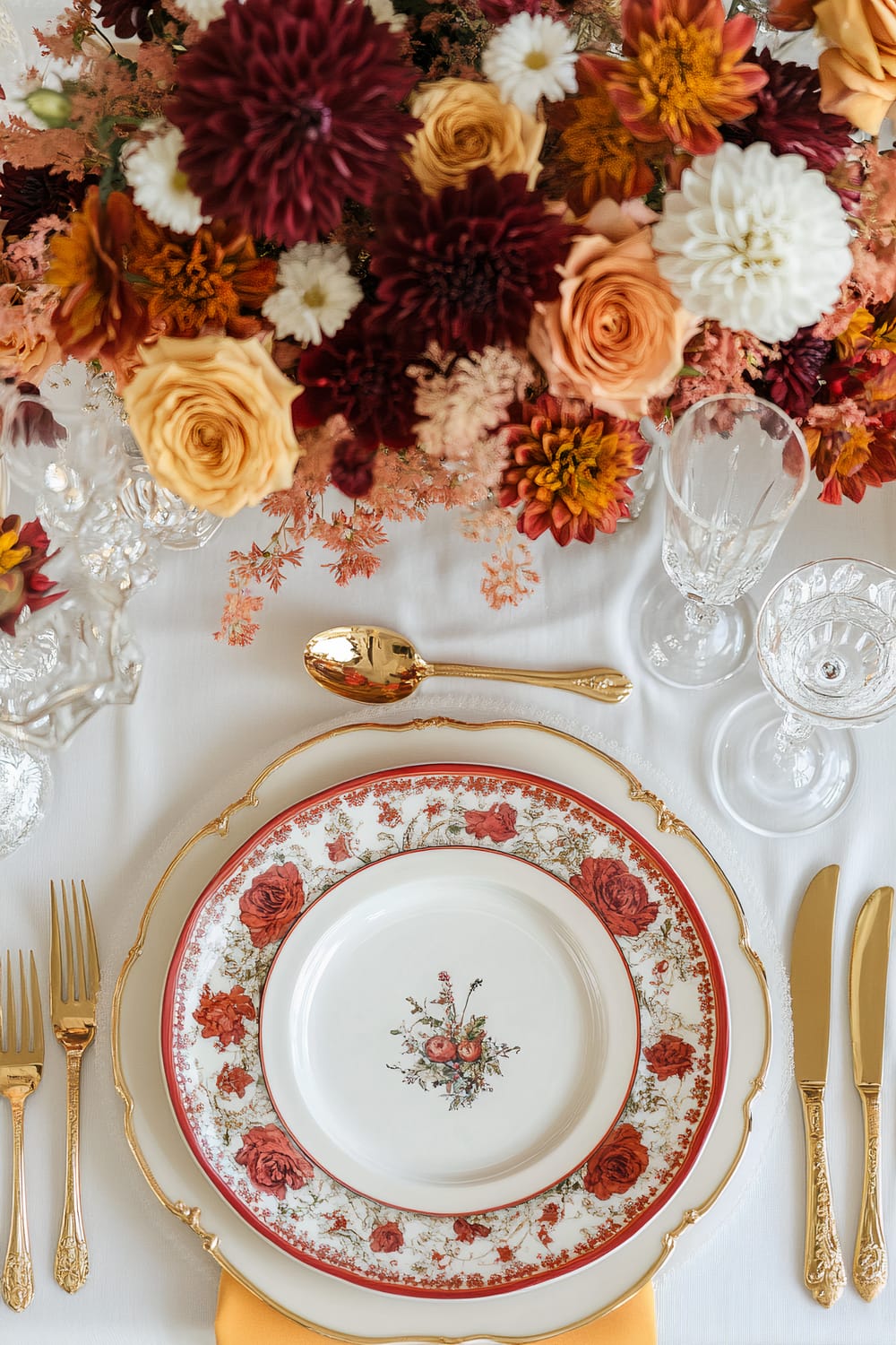 A table setting with elegant floral plates, gold cutlery, and crystal glassware arranged on a white tablecloth. A large, colorful bouquet of roses, chrysanthemums, and other flowers is placed as a centerpiece.