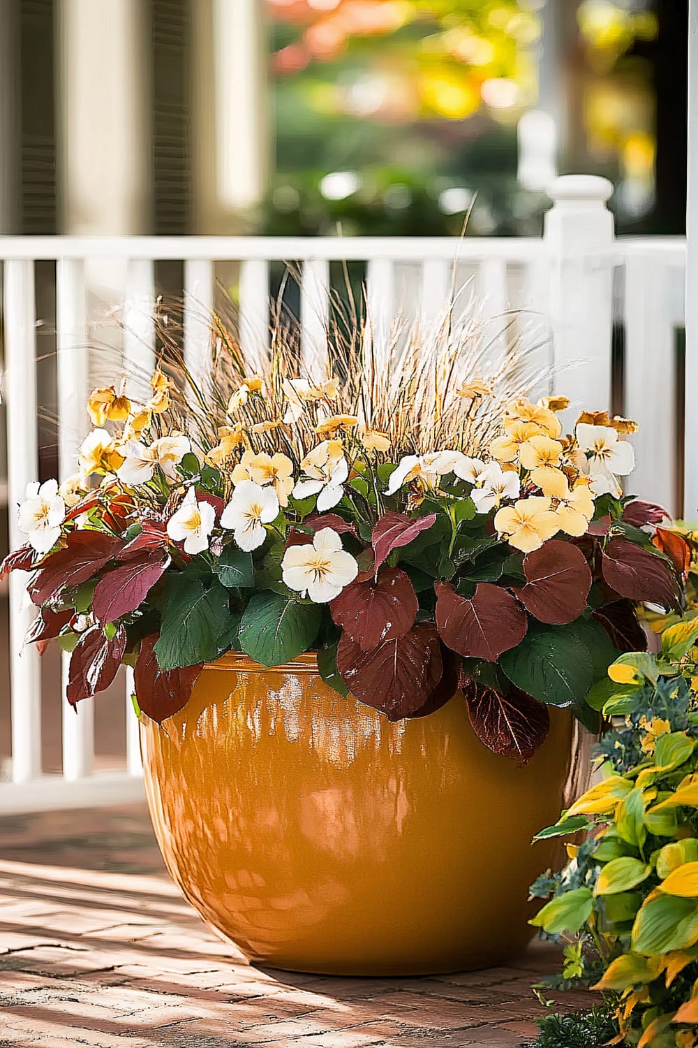 A large, glossy, mustard-yellow ceramic pot sits on a brick patio with a background of white wooden railings. The pot is filled with a lush arrangement of flowers and foliage. The flowers are predominantly soft white and pale yellow pansies, accented by deep burgundy leaves. Tall ornamental grasses emerge from the center, adding height and texture to the arrangement.