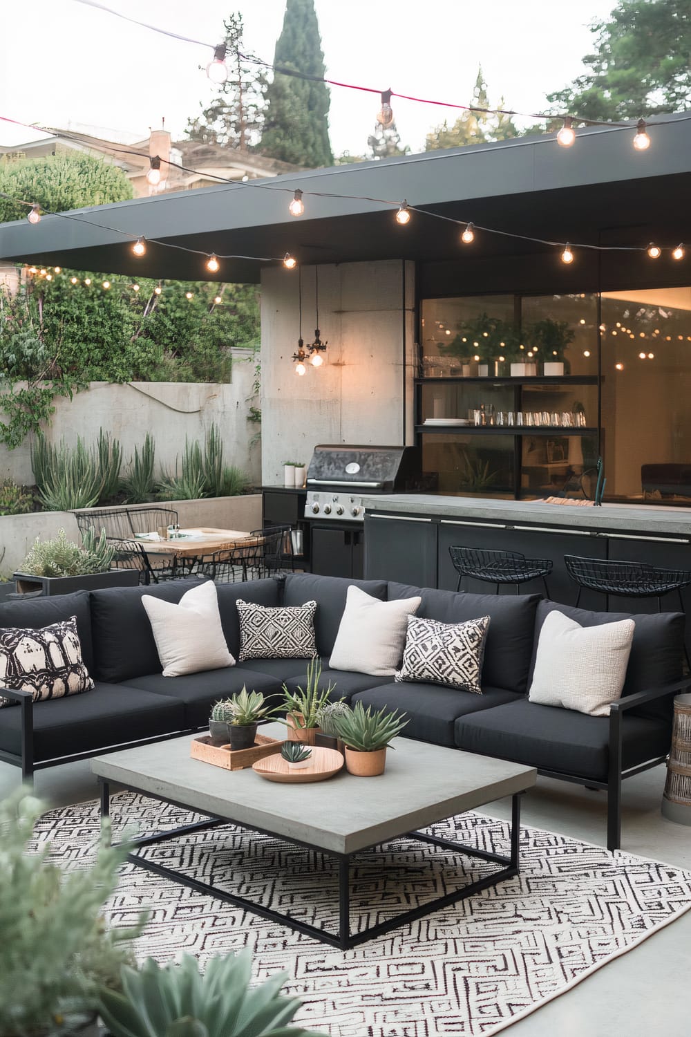 A modern outdoor patio features a sleek black sectional sofa adorned with white and patterned cushions. In front of the sofa, a low, gray coffee table holds several small potted succulents and a wooden tray. The setting is completed with a geometrically patterned rug beneath the coffee table. Above, string lights hang from the patio's overhang, illuminating the area. To the left, there is a dining space with a concrete table and black metal chairs. A built-in grill and bar area are in the background adjacent to a glass wall that provides a view of the well-lit interior.