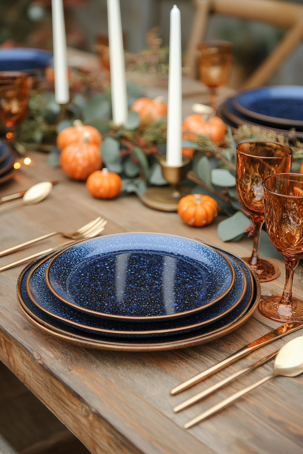 A Thanksgiving dinner table set with sapphire blue ceramic plates, burnt orange glassware, and gold flatware. The centerpiece features white candles on a gold tray, small burnt orange gourds, and deep green foliage. The setting is on a reclaimed wooden table.