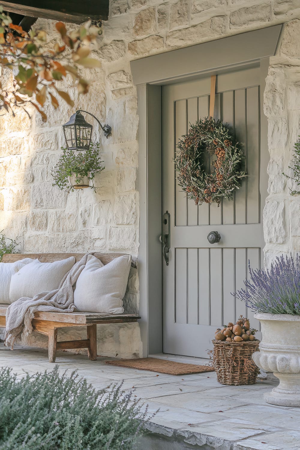 A charming porch scene featuring a stone exterior and a light gray door adorned with a rustic wreath of greenery and pinecones. A welcoming wooden bench to the left is cushioned with soft white pillows and a cozy throw blanket draped over the seat. Hanging beside the door is a black lantern-style light fixture bearing a small potted plant. In the foreground, a basket filled with pears sits on the stone floor next to an earthy, weathered urn containing a lavender plant. Nearby branches with autumn-colored leaves frame the image at the top left corner.