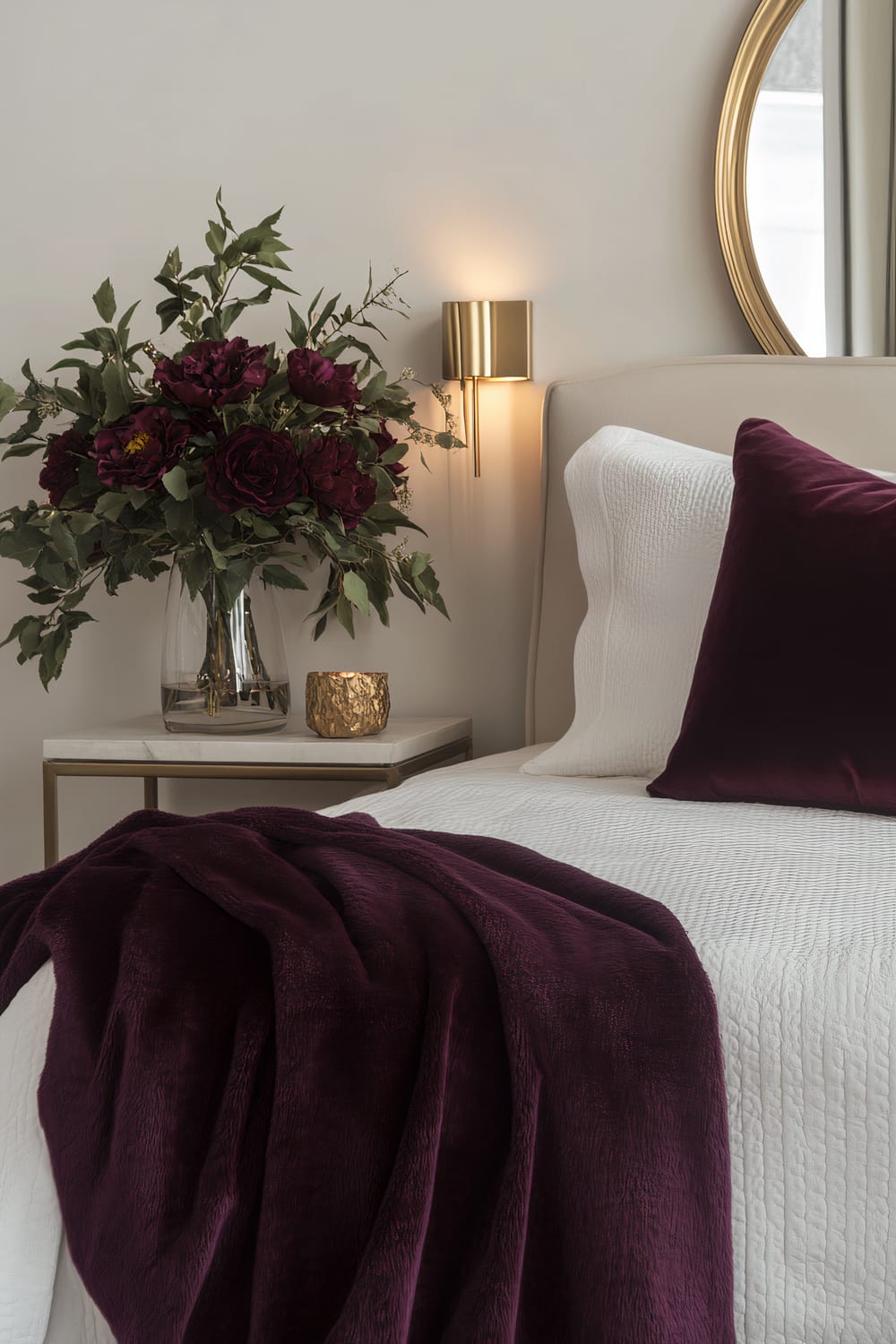 A luxurious bedroom with a thanksgiving theme featuring a deep plum throw draped over a white bed. The bed has white pillows with a single deep plum pillow. A sleek gold bedside lamp and a single large flower arrangement in rich burgundy and deep green hues are on the bedside table. A modern metallic mirror hangs on the wall, reflecting the elegant decor. Soft ambient lighting creates a serene and sophisticated atmosphere.