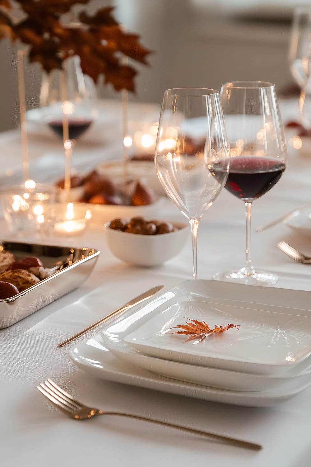 A modern Friendsgiving table setting with minimalist white ceramic plates, vintage-inspired glassware filled with deep red wine, and metallic gold cutlery. The table features a sleek centerpiece with small pumpkins and autumn leaves, elegant serving trays with appetizers, and soft ambient lighting highlighting the sophisticated tableware against a clean white tablecloth.