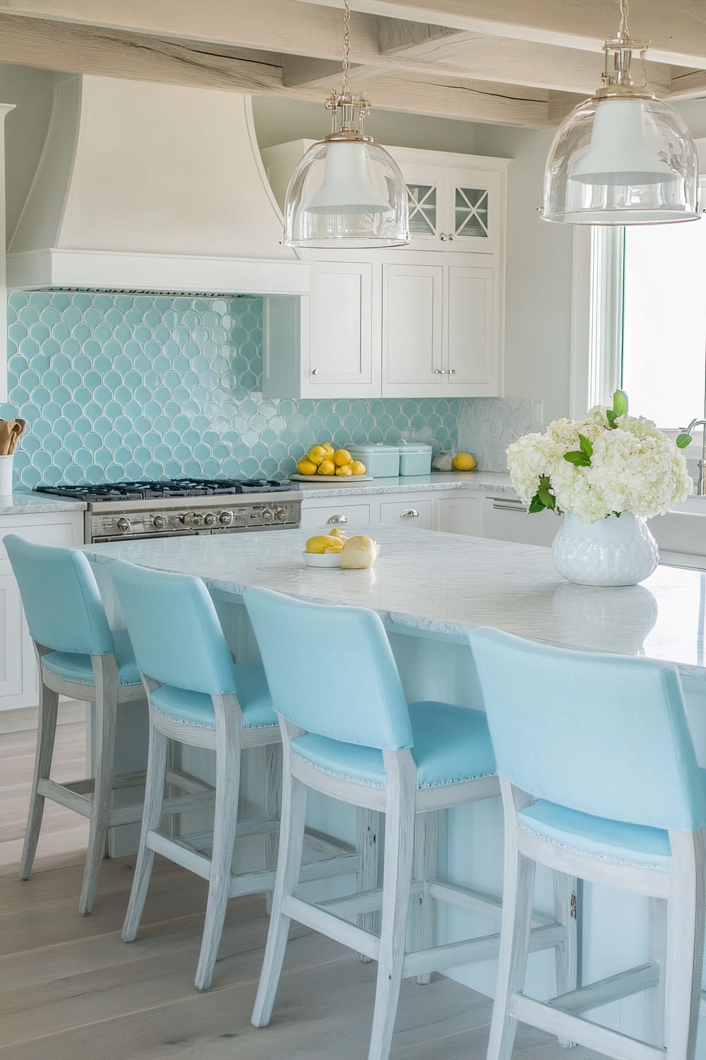 A bright and airy kitchen with a predominantly white and turquoise color scheme. A large white island with a marble countertop and turquoise upholstered bar stools is central to the room. The backsplash features a scallop-patterned tile design in shades of turquoise. Overhead, two large glass pendant lights hang, illuminating the space. There are white cabinets with glass inserts, a stainless steel stove, and a bowl of lemons placed on the counter. A vase of white flowers adds a fresh touch to the island.