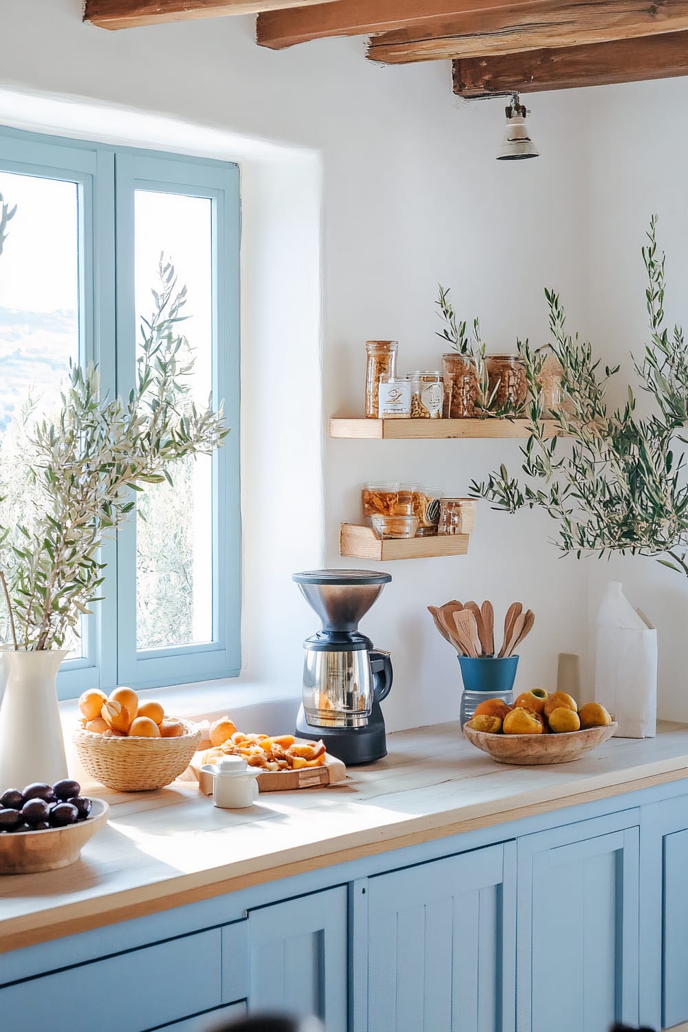 A bright kitchen with blue cabinetry, wooden countertops, and open shelving. The window frames are painted light blue, matching the cabinetry. The countertop has a coffee grinder, bowls of fresh fruits, and a wooden utensil holder. There are olive branches in a vase and shelves holding jars and canisters of dry goods.