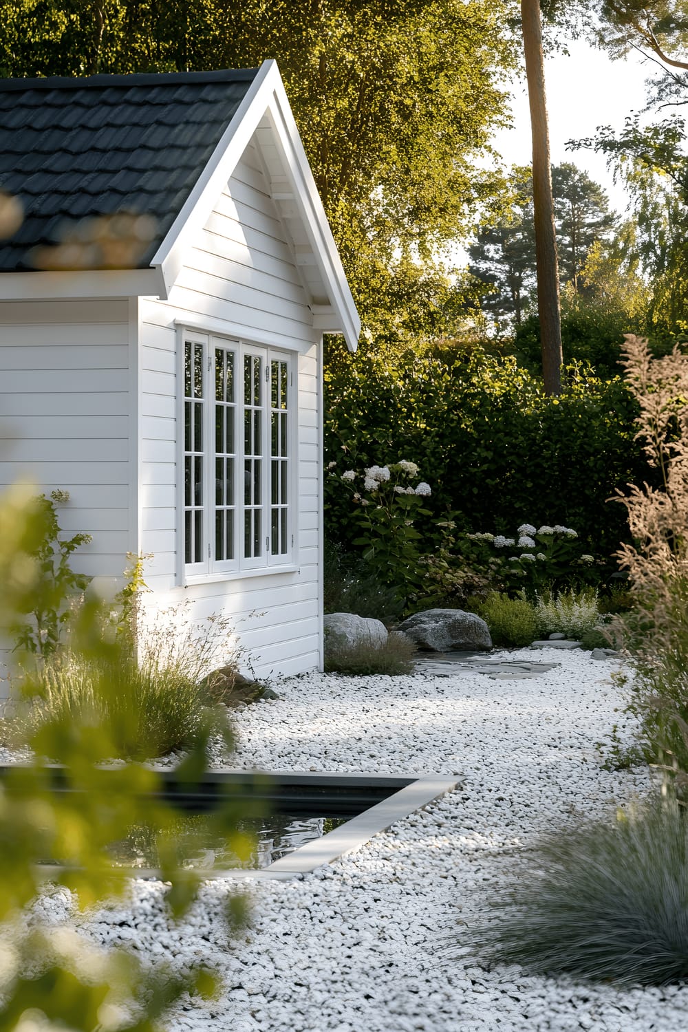 A compact white wooden shed with large windows is set against a backdrop of a sparse garden with light-colored gravel, a few select evergreen shrubs, and a minimalist water feature. The scene is bathed in bright, diffused Nordic daylight, presenting crisp and defined shadows.