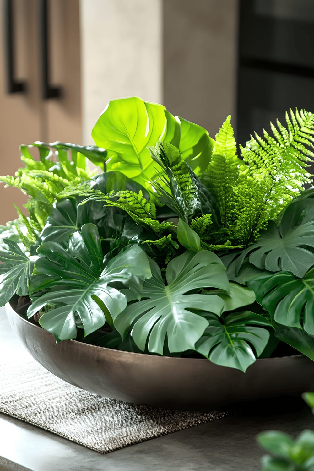 A meticulously arranged industrial-style centerpiece placed on a concrete kitchen table, consisting of a wide, rounded galvanized metal bowl filled with clusters of vibrant green plants, including ferns, pothos, and philodendron. The metallic bowl is surrounded by small metallic geometric shapes and rustic wooden discs, all of which are accentuated by the warm, inviting glow of ambient light. A neutral-toned table runner acts as a base for this harmonious blend of rugged metal elements and the refreshing touch of fresh greenery, providing an intriguing contrast that enhances the overall modern appeal of the kitchen.