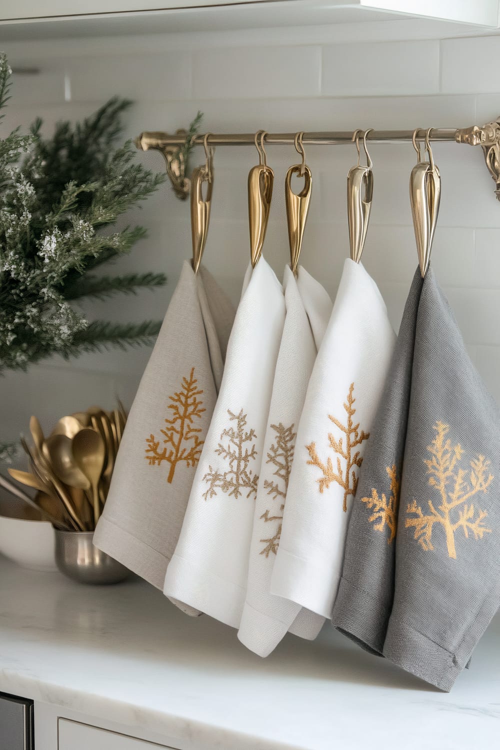 In a modern farmhouse kitchen, five kitchen towels in neutral tones of soft grey, cream, and white hang on gold hooks. The towels showcase subtle gold embroidery in the form of tree designs. Below the towels on the pristine white countertop, a set of stainless steel utensils with gold accents is arranged in a holder. A green plant adds a touch of nature to the scene.