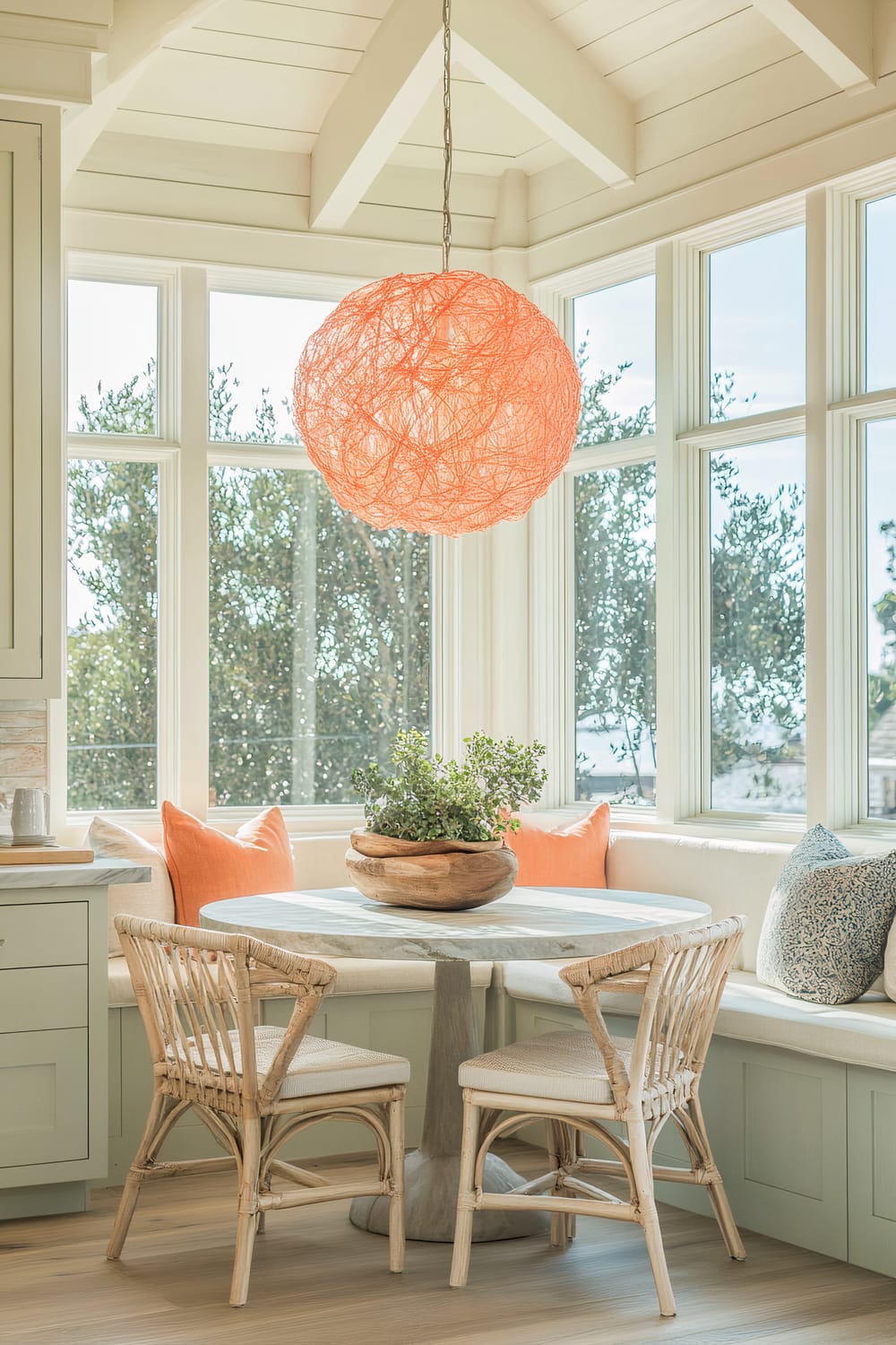 Bright dining nook with a round marble table and wicker chairs, accented by a large orange spherical pendant light, cream built-in seating with bright orange throw pillows, and large windows showcasing an abundance of natural light.