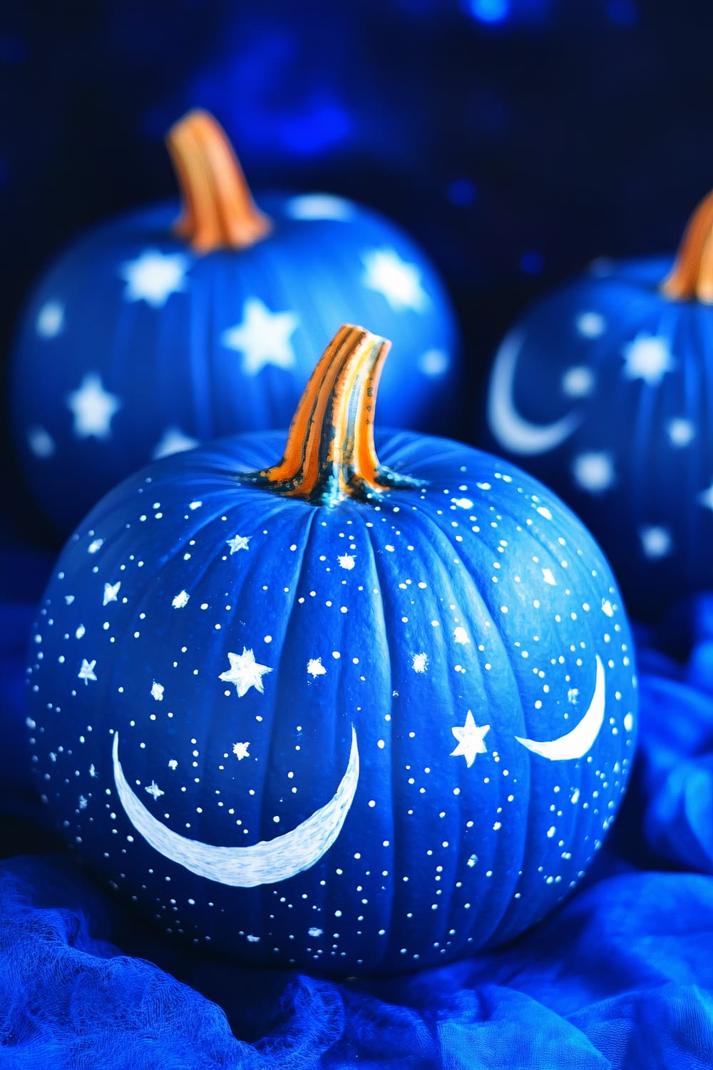 Three pumpkins are painted a dark blue and decorated with white stars and crescent moons, mimicking a night sky. The pumpkins have orange stems and are placed on a blue fabric that adds to the celestial theme.