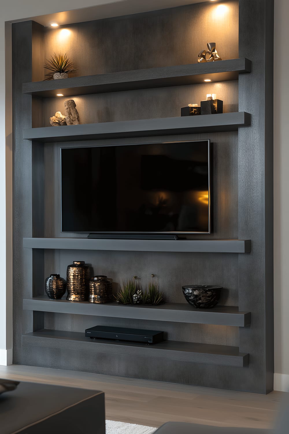 A polished, monochromatic gray TV wall display with multiple stylish floating shelves holding various decor items. The different shades of gray adorning the wall create a sense of depth and subtlety. Among the decor items are matte ceramics and glossy accents, each placed with precision. The whole setup is subtly illuminated by a strip of LEDs integrated into the wall, giving the space a warm and elegant glow.