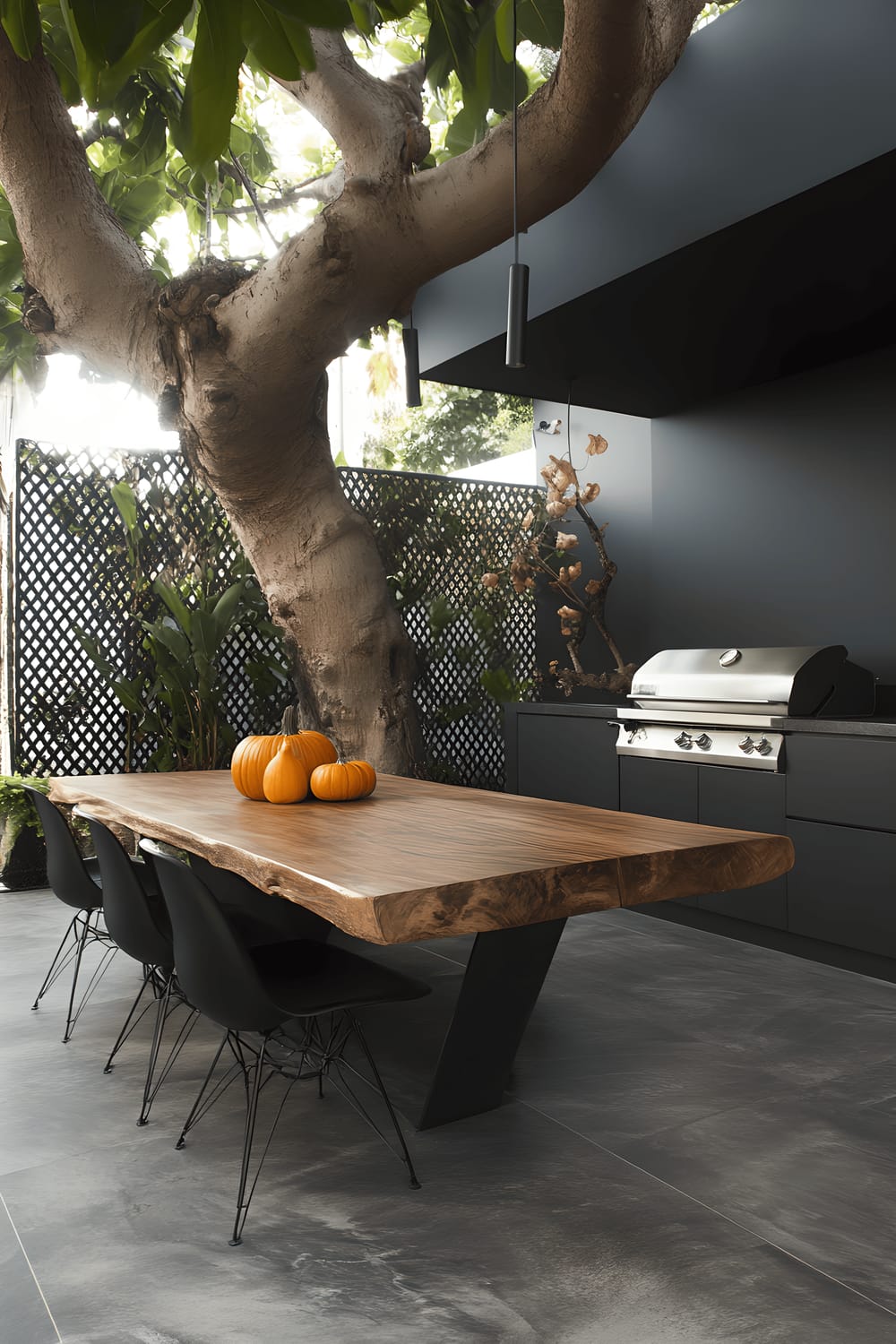 A modern outdoor patio featuring a robust wooden dining table surrounded by sleek black chairs. The grey tiled floor contrasts the vibrant greenery from minimalist planters and the overhead spring tree. ABB lattice privacy walls enclose the area. Finishing touches like a stainless-steel grill and decorative pumpkins contribute to a sense of seasonality and warmth.