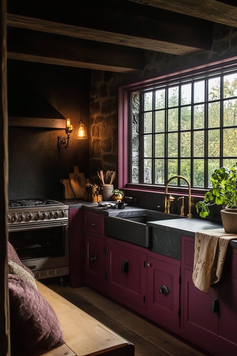 An intimate farmhouse kitchen featuring deep burgundy cabinets and dark slate countertops, viewed from a wooden window seat with cushion. The cabinets are complemented by a stainless steel range and a dark faucet. A large window with black framing allows soft natural light to enter, illuminating the space warmly. The textured reclaimed wood flooring and a stone backsplash enhance the rustic charm of the room.