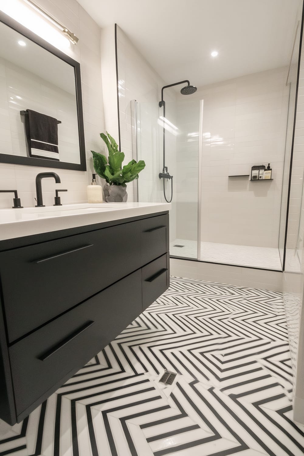 A modern bathroom featuring a black vanity with sleek matte handles and a white countertop. A large mirror with a black frame is mounted above the vanity, reflecting the opposite shower area. The shower has a clear glass enclosure, black shower fixtures, and a small built-in shelf holding toiletries. The floor is covered with bold, black and white herringbone patterned tiles. A green potted plant sits on the counter next to a soap dispenser, adding a touch of freshness to the minimalist design.