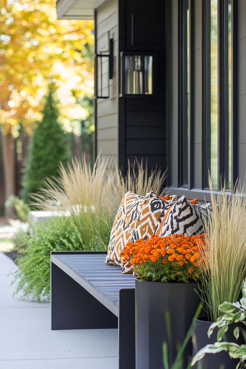 A modern outdoor seating area with a sleek black bench adorned with colorful patterned cushions in orange and white. The bench is flanked by black planters, one of which is filled with vibrant orange flowers, while the surrounding area features lush green foliage and ornamental grasses. The backdrop includes a dark-paneled exterior wall with large windows and a contemporary lantern-style wall light. The scene is set against a backdrop of trees with autumn foliage, blending nature with sophisticated design.