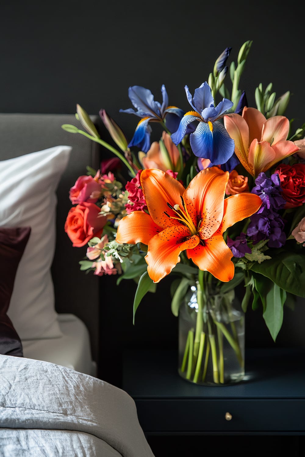 A vibrant bouquet of fresh flowers in a clear vase placed on a dark bedside table next to a bed with white and dark-colored pillows. The bouquet features an assortment of flowers, including a prominent orange lily, blue iris, pink roses, and other colorful blooms against a dark background.