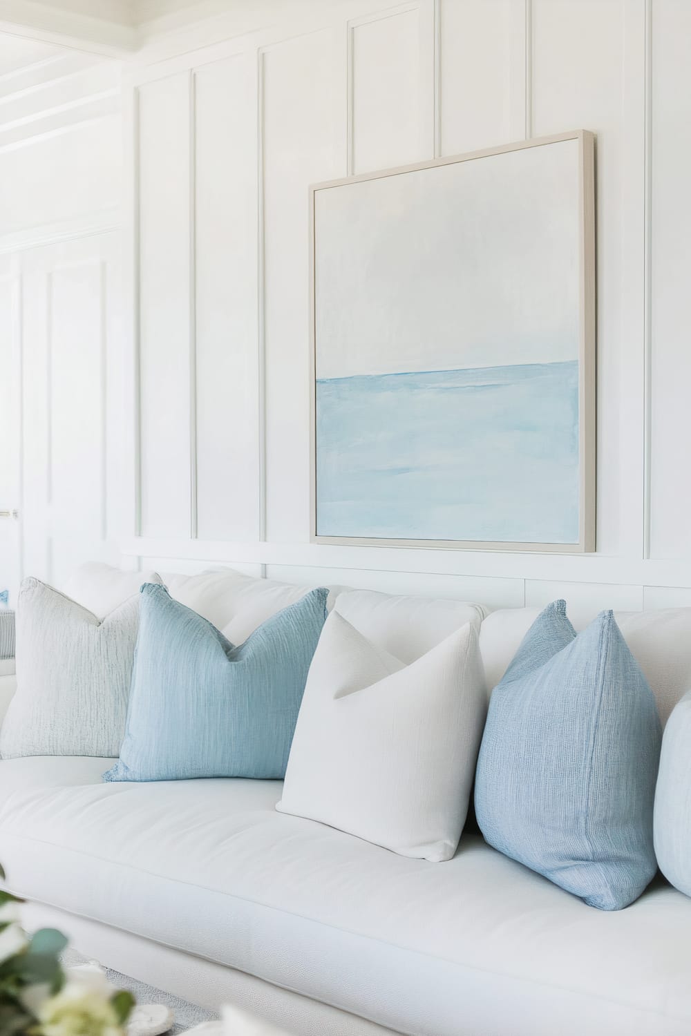 A white sofa adorned with a mix of light blue and white throw pillows is positioned against a white paneled wall. Above the sofa hangs a minimalist painting featuring a serene seascape with a light blue ocean and an off-white sky, framed in a simple light-colored frame. The room exudes a clean and tranquil coastal aesthetic.