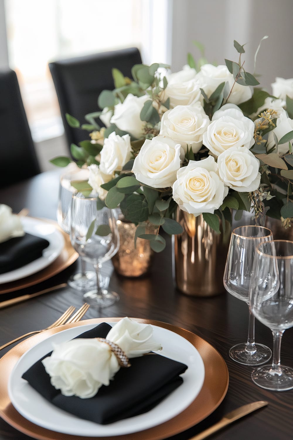 An elegant dining table setup with black chairs. Each place setting includes a white plate topped with a black napkin and a white rose, all placed on a copper charger. Gold cutlery is arranged on either side of the plates. The table centerpiece consists of a bouquet of white roses and greenery in a gold vase, accompanied by wine glasses and smaller gold votive holders.