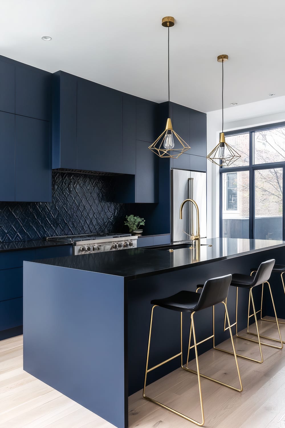 A modern kitchen featuring navy blue cabinetry, black quartz countertops, and gold accents. The backsplash has a diamond pattern in black tiles. A large island with a black surface has gold and black bar stools. Two geometric gold pendant lights with exposed bulbs hang over the island. A stainless steel refrigerator and a gold faucet add a touch of elegance. The kitchen has large windows and light wooden flooring.