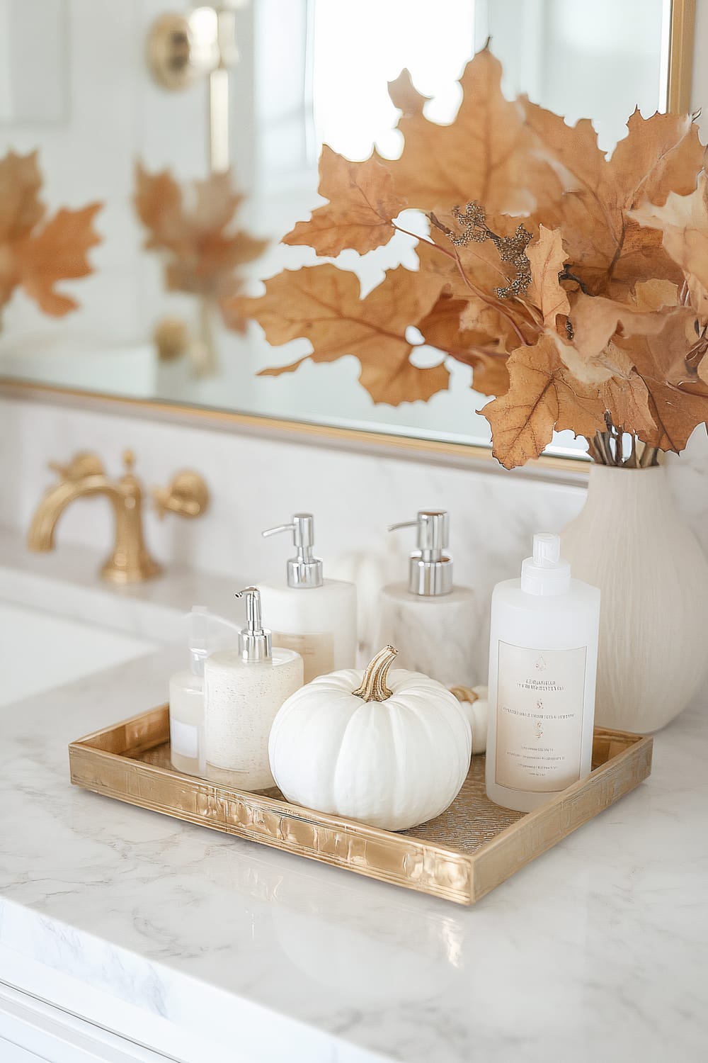 A bathroom vanity featuring a marble countertop with a gold faucet. A gold tray sits on the counter holding various elegantly packaged toiletries and a white pumpkin. A bouquet of dried orange leaves in a cream vase adds a touch of autumn décor, and a gold-framed mirror is seen in the background.