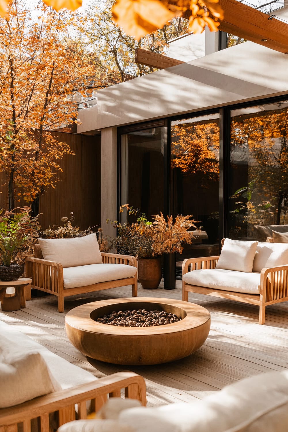 A beautifully designed outdoor patio features natural wooden furniture with white cushions, set around a large, circular wooden fire pit. The patio is surrounded by autumn foliage in shades of orange and yellow, and large glass windows reflect the vibrant colors of the trees.