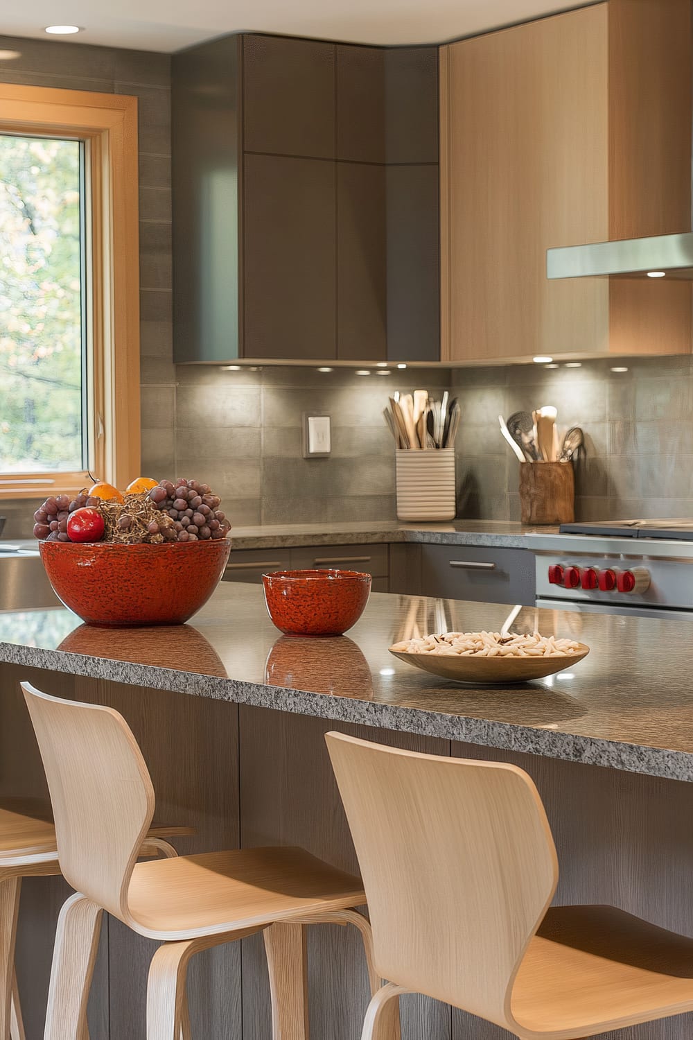 A modern kitchen featuring a granite countertop island with light wood bar stools. The island has a red bowl filled with fruit, a smaller red bowl, and a wooden plate with snacks. The background includes a stove with red dials, sleek cabinetry, and a window with a wooden frame, offering a view of greenery outside.