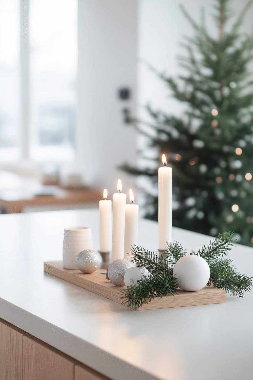 A Scandinavian-style kitchen with light birch wood cabinets and white countertops. A minimalist Christmas display on the kitchen island features a silver candle holder with white pillar candles, surrounded by white ceramic ornaments and a sprig of evergreen. A Christmas tree is visible in the background. Natural light illuminates the space through large windows.